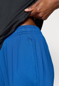 Μπλε αθλητικά παντελόνια με ελαστική μέση και πλαϊνές τσέπες, συνδυασμένα με μαύρο αθλητικό τοπ, ανυψωμένο εν μέρει για να φαίνονται τα παντελόνια.