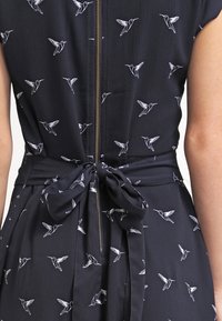 Marinblå klänning med dragkedja baktill, med ett mönster av vita kolibrier och ett knytbälte i midjan, tillverkad av lättviktsmaterial.