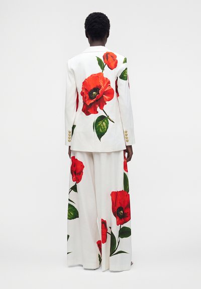 Personne portant un costume blanc avec une grande fleur de coquelicot rouge et un imprimé de feuille verte, vue de dos sur un fond clair uni.