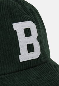 Grønn kordfløyelslue med et hvitt, følt bokstav "B"-patch, med teksturerte ribber og buet skjerm.