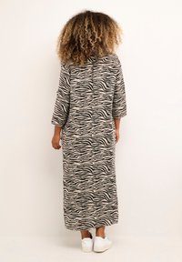 Abito maxi in tessuto di maglia beige con strisce zebrate nere, caratterizzato da maniche a tre quarti e silhouette dritta. Indossato con sneakers bianche.