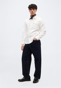 Νεαρός άνδρας φορώντας λευκό πουκάμισο με μακριά μανίκια και κουμπιά, μαύρο μαντήλι γύρω από τον λαιμό, σκούρα φαρδιά τζιν και μαύρα παπούτσια, στέκεται με το ένα χέρι στην τσέπη.