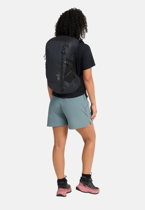 Personne debout de dos portant une chemise noire, un short bleu sarcelle, des chaussures de randonnée et un grand sac à dos noir.