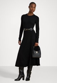 Vestido negro de manga larga, tejido texturizado, ajustado en la cintura con un cinturón negro brillante. Botas altas de cuero negro hasta la rodilla y un bolso pequeño con herrajes dorados.