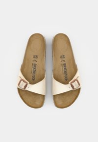 Lys beige sandaler med en glat læderrem, spændeaccent og en brun polstret fodseng. Flad sål og åbent tådesign.