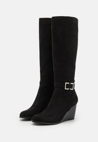 Bottines en daim noir, hauteur genou, à talon compensé, dotées d'un bout arrondi et d'une sangle décorative avec une boucle en argent. Texture lisse dans l'ensemble.