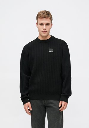 Suéter preto de malha com textura canelada, decote redondo e um pequeno logótipo retangular no peito. Combinado com jeans escuros.