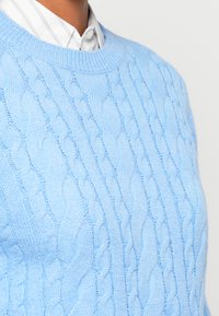 Henkilö, jolla on päällään vaaleansininen palmikkoneule, valkoisen kauluspuseron päällä, jossa on harmaat raidat, kuvattuna kaulasta rintaan.