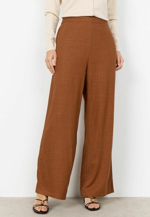 Brązowe spodnie o szerokich nogawkach o średnim stanie, noszone z beżową bluzką na guziki i czarnymi sandałami na paski ze złotymi akcentami, stojące na szarej podłodze.