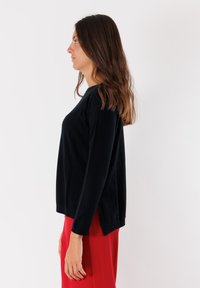 Maglione nero lavorato a maglia con maniche lunghe e una vestibilità rilassata. Fessure laterali notevoli e tessuto testurizzato. Abbinato a un pantalone rosso.