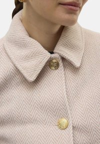 Manteau en laine chevron rose clair avec col, doté de fermetures à boutons dorés et d'une texture lisse. Gros plan montrant les détails des boutons et le motif du tissu.