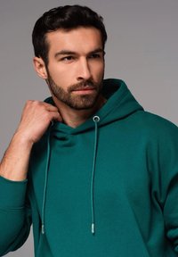 Felpa teal con cappuccio e lacci, dotata di aglet in metallo argento. Il tessuto appare morbido con una vestibilità rilassata e una tasca a canguro.