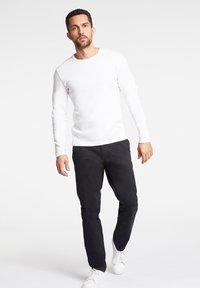Weißes langärmeliges Hemd, kombiniert mit schwarzen Hosen und weißen Sneakern. Das Hemd hat einen runden Halsausschnitt; die Hosen sind schmal zulaufend und strukturiert.