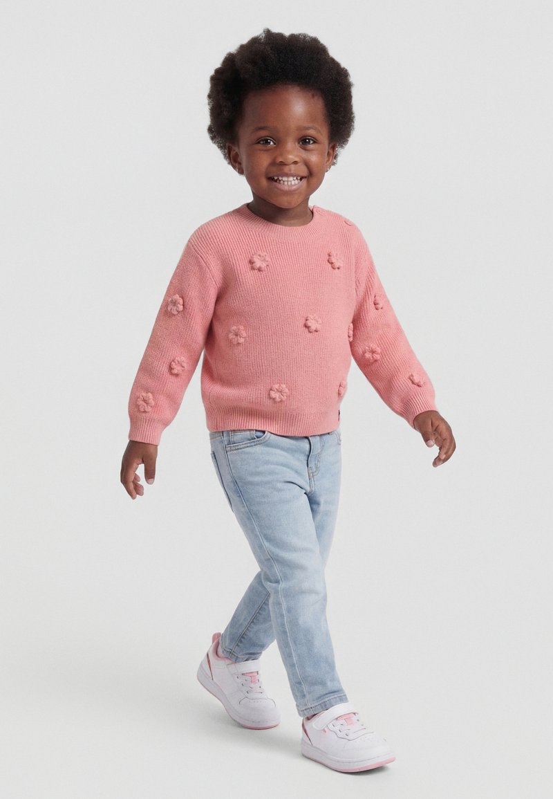Usmievavé malé dieťa kráčajúce, oblečené v ružovom svetri s 3D kvetinovými detailmi, svetlomodrých džínsoch a bielych teniskách s ružovými akcentmi.