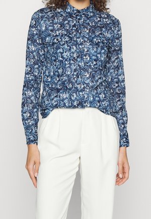 Frau trägt ein blaues, langärmeliges Hemd mit Blumenmuster, das in hoch taillierte weiße Hose gesteckt ist.
