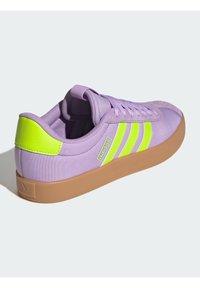 Fioletowy tenisówka na płótnie z trzema limonkowymi paskami, wyściełanym kołnierzem i brązową gumową podeszwą. Charakterystyczna ze względu na teksturowaną powierzchnię i kontrast kolorów.