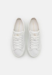 Bílé plátěné tenisky se šněrováním, kulatou špičkou a béžovým logem na jazyku. Hladká textura a minimalistický design.