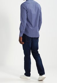 Blå och vit rutmönstrad långärmad skjorta med knäppning på ärmarna, i kombination med mörka denimjeans och vita sneakers, sedd bakifrån.