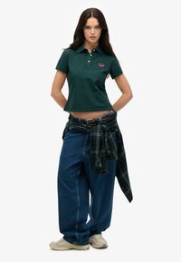 Donkergroen polo met korte mouwen en drie knopen, gecombineerd met losse blauwe denim jeans en een zwart-groen geruit flanelhemd dat om de taille is gebonden.