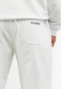 White sweatpants featuring a back pocket with "ICE ICEBERG" embroidered in black. Made of soft, smooth fabric with a relaxed fit.