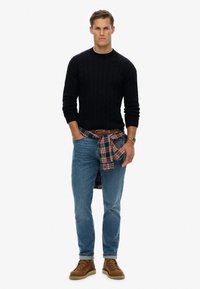 Marineblauwe kabelgebreide trui, lichtblauwe jeans, geruite shirt om de taille geknoopt, bruine laarzen en een horloge met een metalen band.