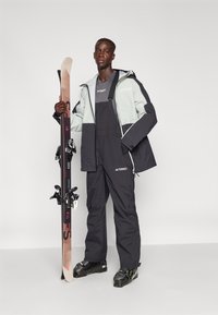 Skidress med en lys grå og svart jakke og matchende overaller. En skikjører holder et par ski med bindinger.