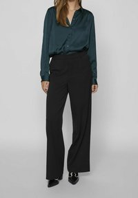 Blouse en satin vert à manches longues et boutons, associée à un pantalon large noir. Chaussures en cuir verni noir avec des talons sculptés.