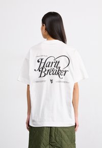 Bílý bavlněný tričko s velkým černým grafickým potiskem na zadní straně, s textem "Hartt Breaker" a uvolněným střihem.