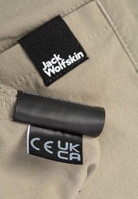 Etichetta in tessuto beige chiaro con logo nero "Jack Wolfskin" e etichetta grigia "CE UK", cucita su materiale testurizzato.