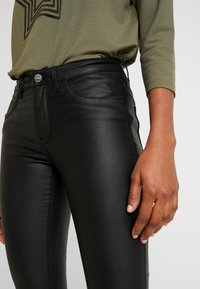Pantalones ajustados de imitación de cuero negros con una textura suave, que presentan un diseño de cinco bolsillos y un cierre de un solo botón. Llevados con una blusa verde oliva.