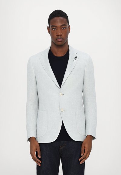Blazer gris clair en tissu texturé, avec deux boutons, des revers crantés et deux poches avant. Porté par-dessus une chemise sombre.