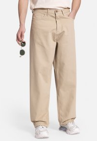 Pantalones de algodón beige con un corte relajado, bolsillos frontales y una textura ligeramente descolorida. Acompañados de gafas de sol redondas y reflectantes.