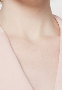 Světle růžové oblečení s dekoltem do V, ukazující hladkou tkaninu a jemnou texturu, zdůrazňující neutrální tón pleti pod ním.