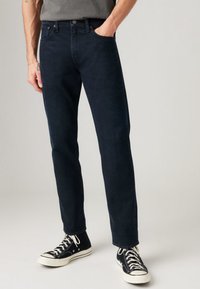 Dark blue denim jeans with a straight leg fit, featuring five pockets and a button closure. Worn with black high-top sneakers.