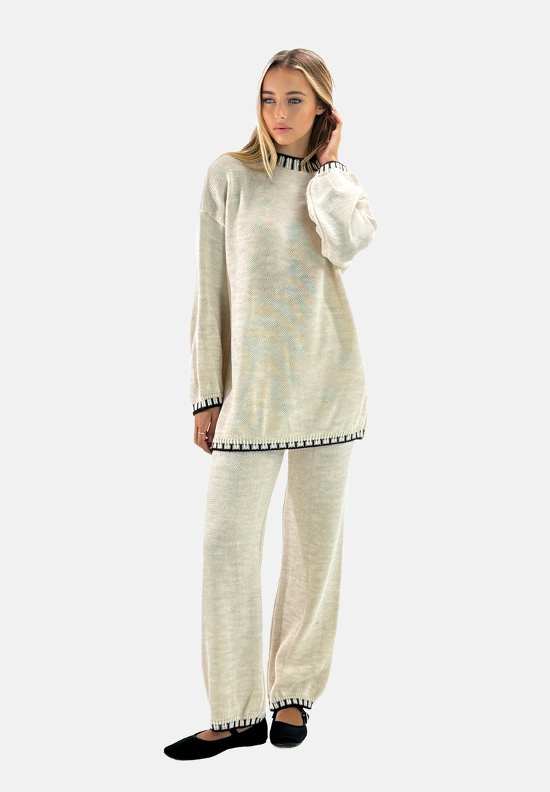 Túnica de punto color beige claro con ribetes negros y mangas anchas, acompañada de pantalones sueltos a juego. Unos zapatos planos negros completan el atuendo.