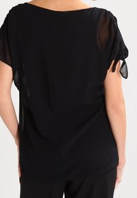 Blusa negra con mangas cortas y transparentes, de corte holgado. Presenta un escote en la parte posterior en forma de pala y detalles fruncidos en los extremos de las mangas.