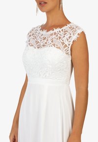 Bílé šaty s krajkovým živůtkem s květinovými vzory, průhledným výstřihem a hladkou splývavou sukní. Krátké rukávy a přiléhavý pas.