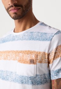 Mann som har på seg en hvit T-skjorte med rund hals, brede blå og orange teksturerte striper og en liten brystlomme med etikett.