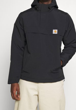 Person iført en sort hættejakke med et Carhartt-logo på brystet og lysebeige bukser, stående foran en ensfarvet baggrund.