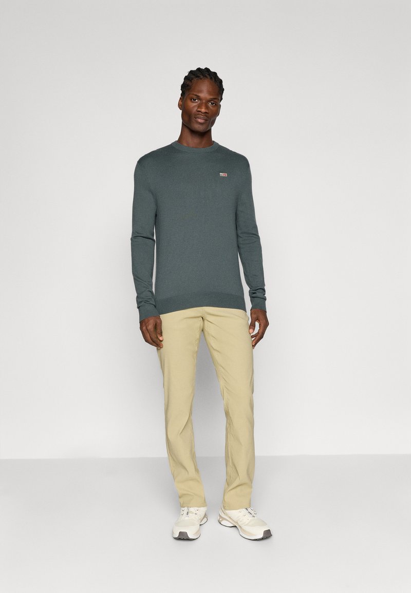 Pull à manches longues vert foncé avec un col rond et un petit logo, associé à un pantalon beige et des baskets blanches.