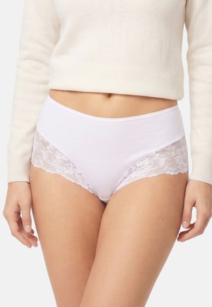 Mujer con braguitas blancas de cintura alta con detalles de encaje floral en los laterales y un jersey corto crema.