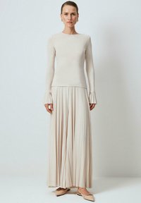 Žena nosi béžový top s dlhými rukávmi a skladanými manžetami, zodpovedajúcu skladanú maxi sukňu a béžové ploché topánky so špicatou špičkou.