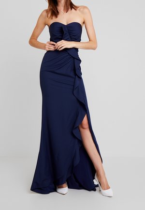 Nainen, joka on pukeutunut olkaimettomaan tummansiniseen mekkoon, jossa on rypytetty yksityiskohta ja reiden halkio, yhdistettynä valkoisiin korkeakorkoisiin kenkiin, seisoo yksiväristä taustaa vasten.