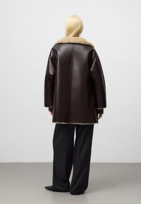 Cappotto in pelle marrone con un morbido collo in pelliccia beige, design oversize, e un maglione di lana scura a contrasto sotto, abbinato a pantaloni neri larghi.