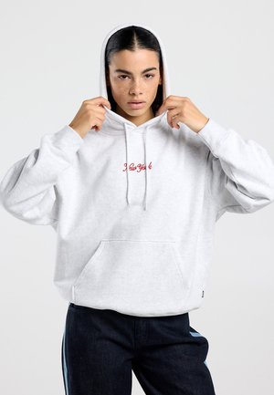 Jongere met donker haar en sproeten, gekleed in een witte "New York" hoodie en een donkere broek, trekt de hoodie over het hoofd.