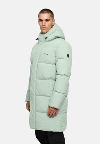 Uomo che indossa un cappotto invernale imbottito verde chiaro con cappuccio, che sta in posizione laterale, su uno sfondo bianco semplice.