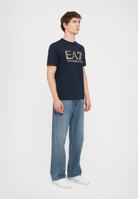 Jūrinis trumpomis rankovėmis marškinėliai su auksiniu "EA7 Emporio Armani" logotipu, derinami su šviesiai mėlynomis plačiomis kelnėmis ir baltomis sportinėmis bateliais.