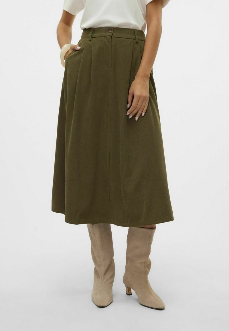 Femme en jupe midi vert olive, haut blanc, bottes beiges montantes, une main dans la poche sur fond uni.