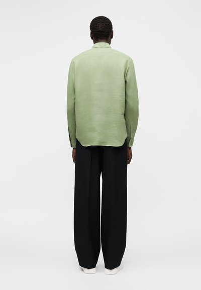 Άνδρας που στέκεται με την πλάτη, φοράει ανοιχτοπράσινο πουκάμισο με μακριά μανίκια, μαύρο παντελόνι με φαρδιά πόδια και άσπρα παπούτσια σε απλό φόντο.