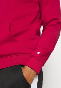 Felpa rossa con tasca a forma di canguro e polsini a coste, con dettaglio logo bianco sulla manica. Indossata con pantaloni neri.